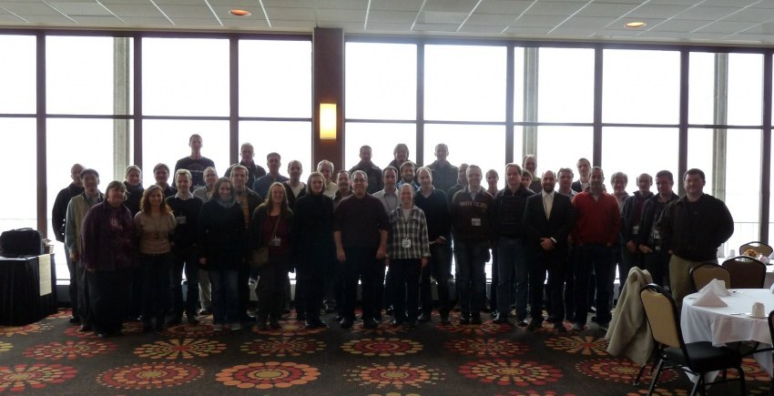 Group photo of about fifty workshop participants standing in front of a large window front